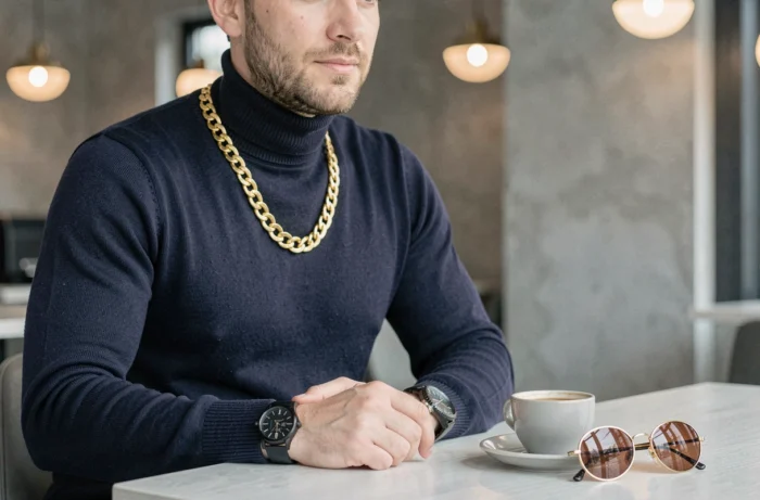 Homme tendance attablé dans un café moderne, portant un col roulé marine, une chaîne dorée et une montre élégante